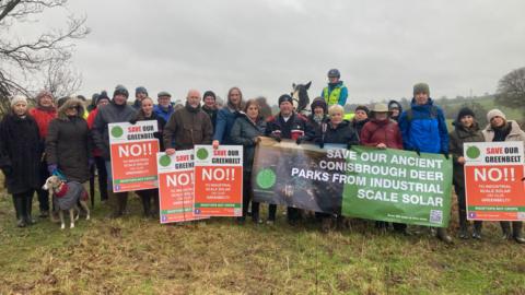 Twenty six people on foot and one horse-rider are in a field, standing in a line three people deep looking at the camera. They are holding banners opposing development on greenbelt land. The largest banner says "Save our ancient Conisbrough deer parks from industrial scale solar" 