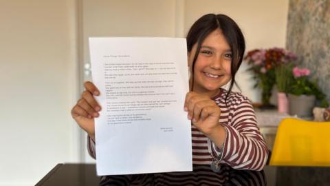 A girl is sat at a kitchen table and is holding up a sheet of paper with a poem written on it.