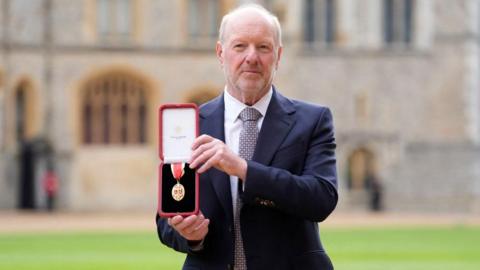 Alan Bates, who wears a navy suit and patterned tie, holds up a gold and red medal, in a red presentation box, outside Windsor Castle