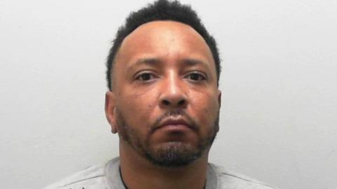 A custody photo of a man wearing a light grey sweatshirt who sits against a plain, pale background, facing forward.