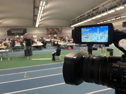 A TV camera in focus with blurred background of vote counters.