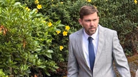 Lewis Baker has dark hair and a dark beard and is walking in front of green bushes. He is wearing a grey jacket, white shirt and blue tie.