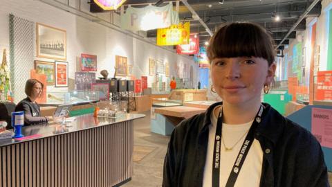 Communications manager Dr Áine McKenny who's wearing a lanyard, blue shirt and white T-shirt underneath, stands in the reception area of the museum's new base in Salts Mill