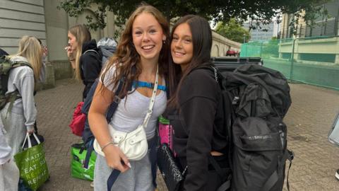 Two smiling young women with large backpacks pose for a photo while waiting in line, surrounded by other festival-goers with camping gear.