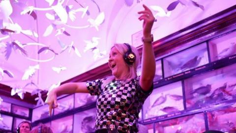 A blonde-haired woman smiles as she dances in an illuminated room. She is wearing headphones.