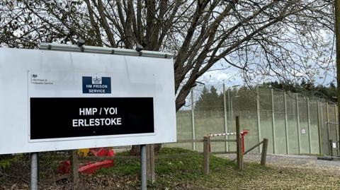 Image of a large road sign for HMP Erlestoke. In the background can be seen high metal security fencing with rolls of barbed wire on the top.