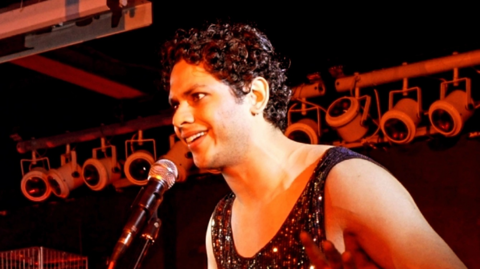Vyome Singh is talking into microphone, which is being held up on a stand, and is standing in front of theatre lights, which are all turned off.