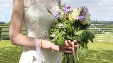 Bride in a cream dress holds a wilting bouquet of summer flowers. She stands in a field with a paddock behind her. 