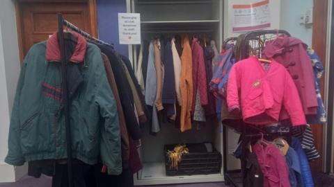 Several coats on a rack. It's on the side of the room in a library. There's a bright pink small coat on one side and a big green coat on the other side.