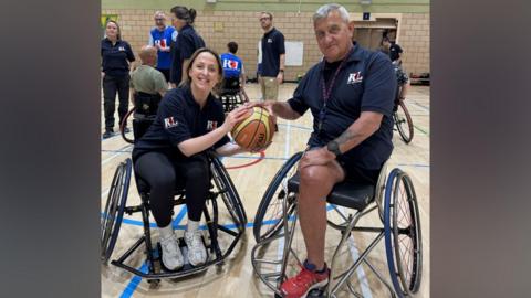 Natalie has light brown hair in a ponytail and she is sitting in a basketball wheelchair, holding onto one part of a basketball. On the other side is a man who is an amputee and has one let - he is sitting in a basketball wheelchair and has his hand on the ball too