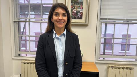 Advocate Natasha Newell. Partner at No.10 Advocates wearing a blue shirt and blazer. She is standing in her office in front of a window.