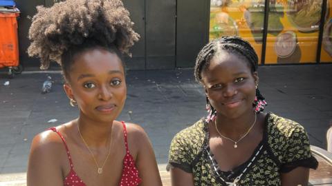 Ms Barumire and Ms Amusat are pictured together on a bench on a sunny day in Manchester. They are both smiling at the camera as the sun shines on their faces. Ms Barumire has her brown hair up and is wearing gold earrings, a gold necklace and a red and white dotted strappy summer dress. Ms Amusat is also wearing gold jewelry with a gold nose ring. She has dark hair in dreads and is wearing a black and yellow top with short sleeves. 