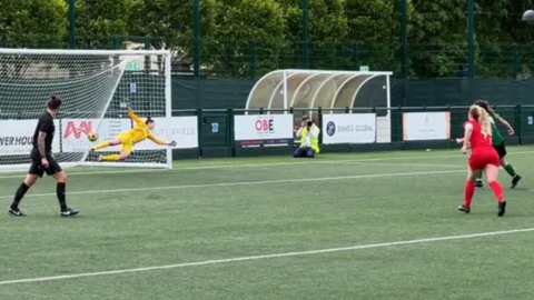 A group of women, one team in red the other in green, playing football - the goal keeper in yellow is diving to their left to save a ball which has gone out of play.