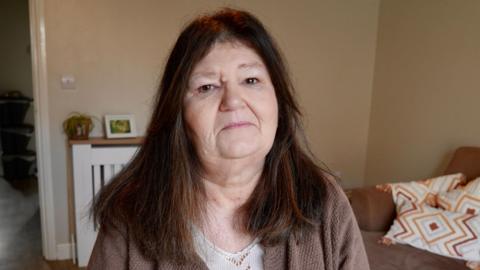Andrea, who has brown long hair and wears a brown cardigan, sitting in her living room with a neutral expression