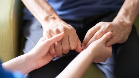 A close up of two people holding hands, one pair of hands visibly older than the other. The two people appear to be sitting down and wearing dark colours.