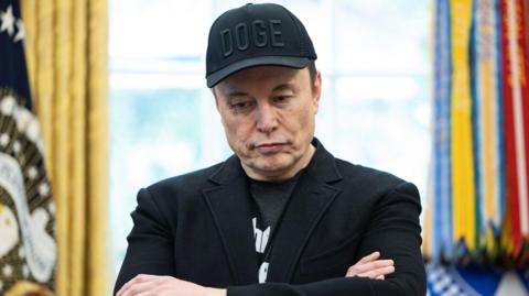 Elon Musk stands in the oval office in a black suit jacket, a black tshirt and a black cap with the letters DOGE stitched in. His arms are folded and he is looking downwards. In the background the flag with the President's standard can be seen to the right, it is out of focus.