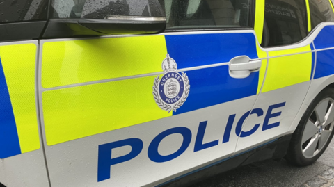 The side of a parked Guernsey Police vehicle. Blue and yellow hi-vis sticks are on the side of the car. A Guernsey Police logo is displayed.
