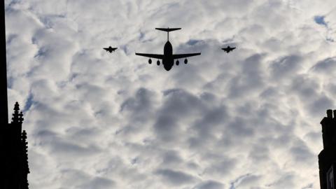 A large cargo plane in silhouette flanked by two of the Red Arrows against a cloudy sky