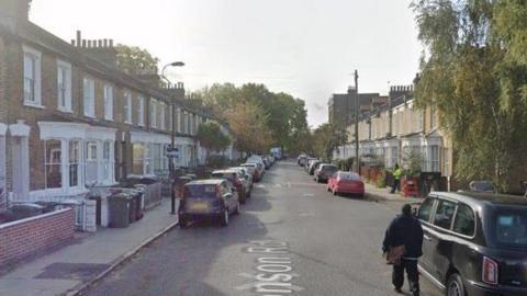 Monson Road in New Cross. A row of light-bricked terraced houses and cars including a black Taxi are on either side.