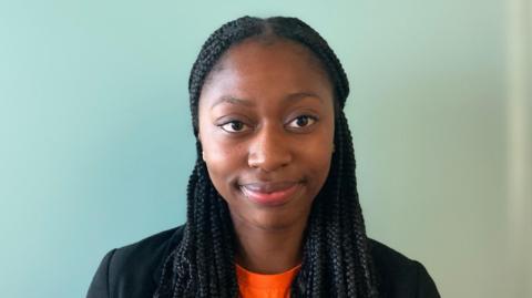 A girl with long black braids smiling, wearing an orange t-shirt and black blazer. She is inside with a wall behind her.