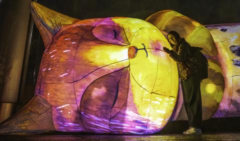 Image shows a large illuminated pink and yellow lantern of a cat in the shape of a cat sleeping on its side. A woman wearing a jacket and Nike trainers is hugging the lantern and smiling to the camera.