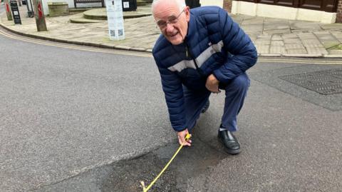 Market Drayton mayor, Roy Aldcroft on the high street, kneeling in the road measuring a 3x2 foot pothole which is 7 inches deep.  Roy is looking at the camera as he is kneeling.