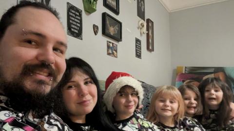 Rowan Penny-Lee with her husband Raven and their four daughters. They are all sat on a sofa wearing matching pyjamas.