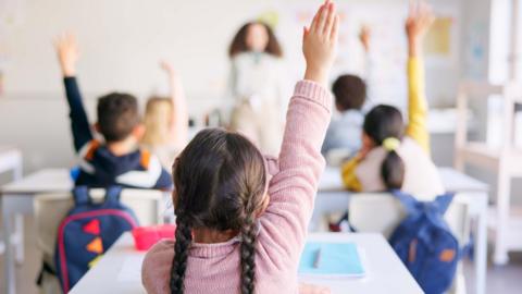 A shot of primary school-aged children from the back raising their hands as they are asked a question by a teacher