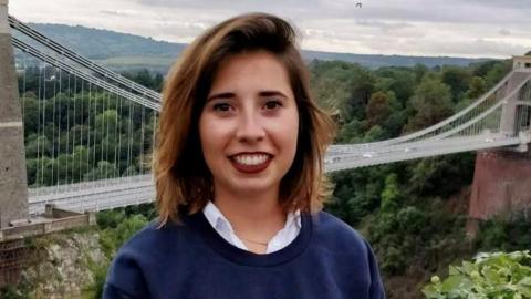 Marta Elena Vento stands with Clifton Suspension Bridge in the background. She has shoulder-length, brown hair and wears make-up, a white shirt and a dark blue top.