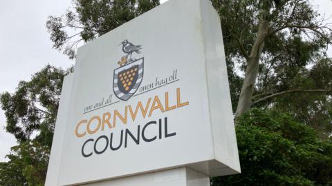 White Cornwall Council sign with the Cornish emblem on it and Cornwall Council written in block capitals. There are trees behind the sign