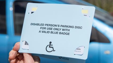 A Blue Badge pass being held up with a blue car in the background. The text on the pass reads: 'DISABLED PERSON'S PARKING DISK FOR USE ONLY WITH A VALID BLUE BADGE'.