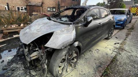 A burnt-out car in front of terraced houses, with its charred engine fully exposed