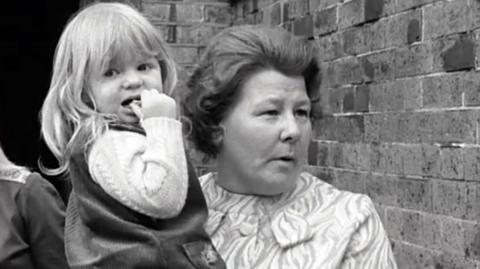 A lady speaking to the interviewer, holding a child.