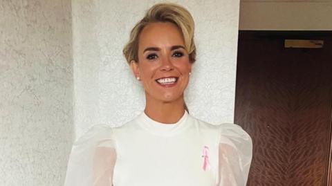 A headshot of a woman smiling into the camera. She is wearing a white dress with puffy, sheer sleeves and has a pink breast cancer ribbon pinned to the right-hand side of her dress. She has blonde hair which is in an up-do and is wearing pearl earrings. 