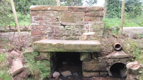 An ageing brick and stone culvert spanning a fast-flowing stream. On the muddy banks to either side are leafless trees, with more in the background alongside a narrow road.