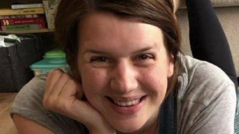 Jennifer Cahill with long brown hair wearing a grey t-shirt in a home with books on a bookshelf behind her in the background. She is smiling.