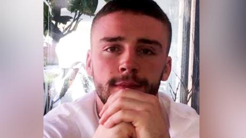 A young man with short, dark hair and stubble looks into the camera with his hands clasped in front of his chin. He is wearing a white T-shirt and is sitting in front of a glass pane.