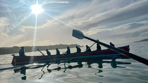 A side-on image of a crew sat in a gig with oars out of the water. 