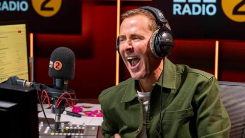 Scott Mills in his Radio 2 studio. He's sitting in front of a microphone with a wide smile, like he's laughing
