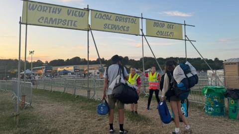 Two people leaving Glastonbury Festival. They are carrying lots of luggage and belongings on their backs. Two stewards in high vis vests are pointing to direct them out of the site. There are three designated queues fenced off for Worthy View Campsite, local/Bristol Services and the drop off/collect area.