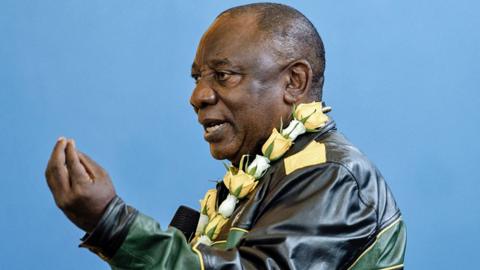 Cyril Ramaphosa, wearing a leather green, yellow and black leather jacket and with a yellow and white rose garland around his neck, gestures with his hand as he talks.