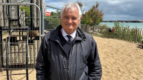 A man called Mark Labey stands in a play park on a seafront. He is wearing a black coat, black jumper, white shirt and black tie. He has trimmed white hair. The park's play equipment is fenced off. The park has sand on the floor.
