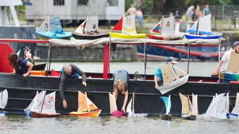 Five people leaning over the edge of a black river boat, lowering colourful handmade sailboats into Bristol's harbour. There are more model boats resting on the canvas roof of the boat. 