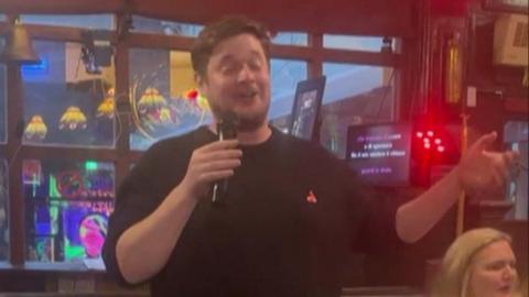 Man in black sweater closes his eyes as he sings into microphone - a woman in a white shirt with blonde hair sits behind him. You can see the words on karaoke machine.