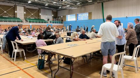 People standing around tables in a sports hall counting votes