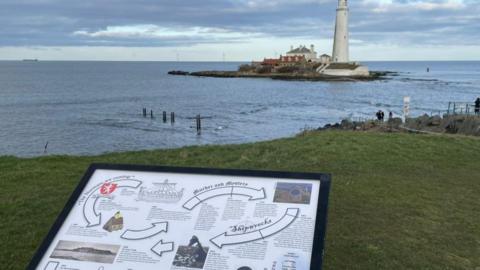 The foreground of the photograph shows a visitor information board with St Mary's Lighhouse in the distance across a stretch of water.