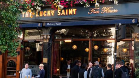 A pub with punters outside covered in flowers and woodwork. Many people stand outside with pints.