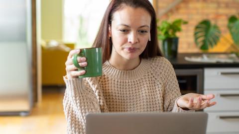 A young woman holds a green mug and looks exasperated at an open laptop