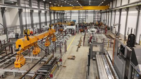 View of a factory floor, with crane-like machinery to the left and a conveyor belt to the right. Further machines can be seen stretching into the distance