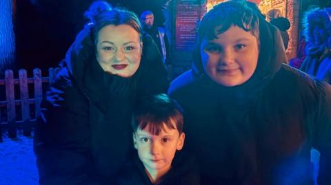 A photo of three people. A woman is on the left smiling and has a black puffa coat on, a boy is to her right and also has a black puffa coat on and a younger boy is in the middle looking at the camera with short brown hair. 